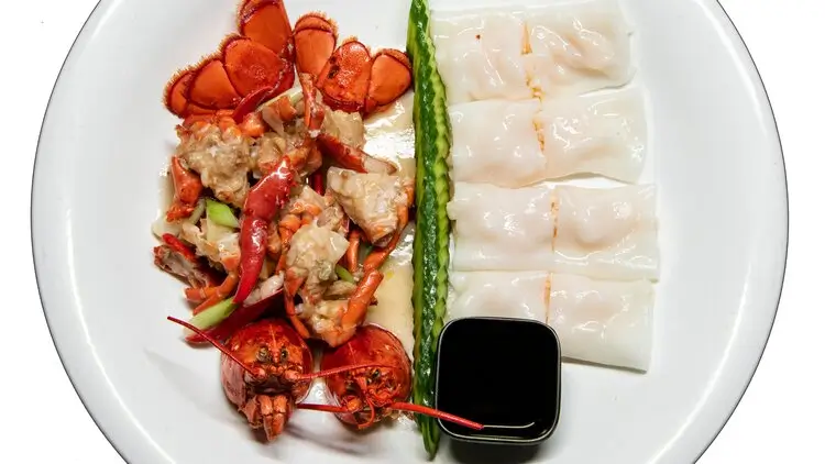 A gourmet dish featuring lobster pieces and shrimp dumplings is arranged on a white plate. A single asparagus spear separates the two main components, and a small dish of soy sauce is placed near the dumplings.